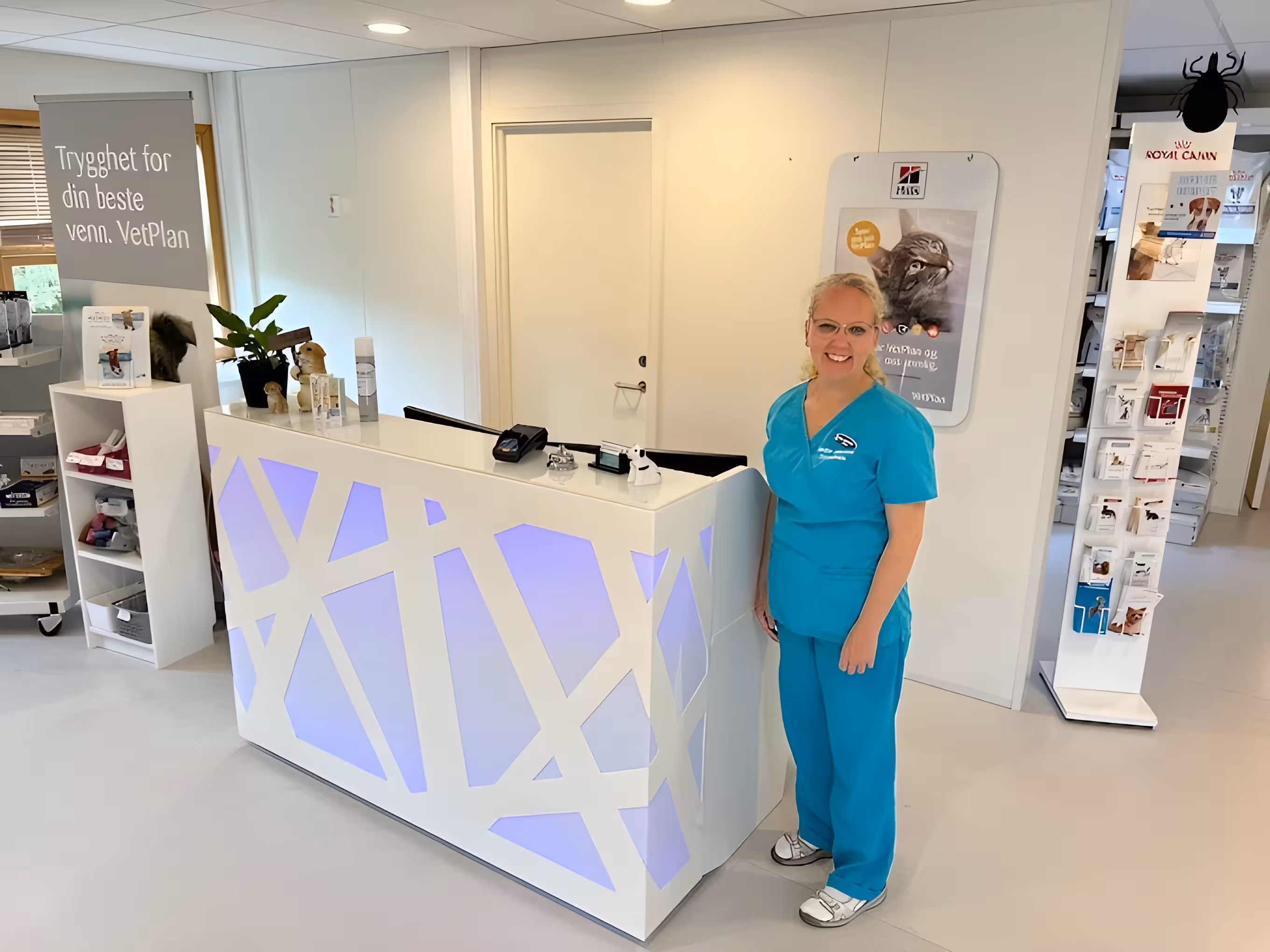 Veterinær i turkise arbeidsklær står smilende ved resepsjonsdisken i en lys og ryddig klinikk, med informasjonsbrosjyrer, betalingsterminal og reklamemateriell i bakgrunnen.